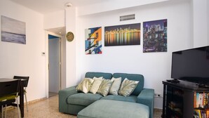 Ein Wohnzimmer mit einem grünen Sofa, einem Esstisch mit Stühlen, einem Bücherregal und drei Bildern an der Wand.