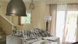 Ein Wohnzimmer mit einer Couch, einem Couchtisch und einem Fernseher. Es gibt ein Fenster mit Vorhängen und eine Lampe.