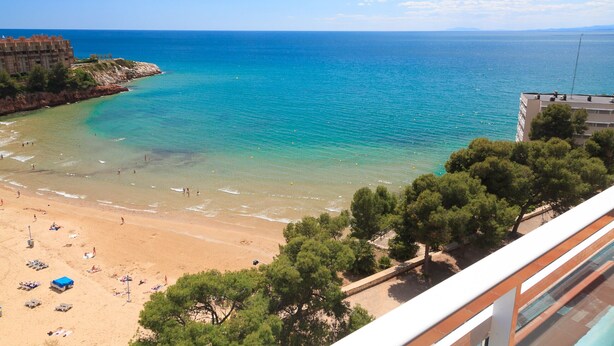 Ein Strand mit klarem türkisblauem Wasser, einem sandigen Ufer und einem Gebäude mit einem Balkon mit Blick auf das Meer.