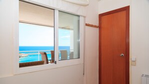Ein Zimmer mit Fensterblick auf einen Strand, einer Holztür und einer kleinen Sitzecke mit Stuhl und Tisch.