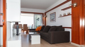 Ein modernes Wohnzimmer mit einer Couch, einem Couchtisch und einem Fernseher. Ein Essbereich mit Stühlen und einem Tisch ist durch eine Glastür sichtbar.