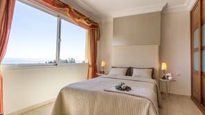 A hotel room with a large bed, two bedside lamps, a view of the sea, and a window with curtains.