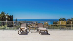 A balcony with two lounge chairs and a small table, overlooking a coastal view.