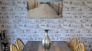 A dining table with six wooden chairs, a metal vase, and a beach-themed painting on a brick wall. - Gower, Oxwich, Amazing Views, Sleeps 4 (Oxwich)