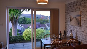 A dining area with a table set for four, a sliding glass door leading to a garden, and a brick wall with a framed picture. - Gower, Oxwich, Amazing Views, Sleeps 4 (Oxwich)
