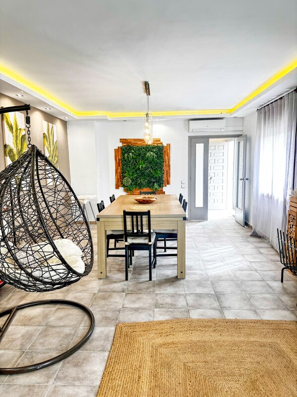 A dining area with a wooden table, chairs, a hanging chair, a green wall panel, and a rug on the floor.