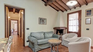 Una sala de estar con sofá, una mesita y una chimenea. Hay una puerta que conduce a otra habitación y una ventana con cortinas.