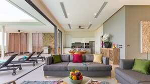 A modern living room with a grey sofa, a coffee table with magazines, and a balcony with sun loungers. - Baan Ouyan 5-bed luxury family home on Bophut Hill (Ko Samui)