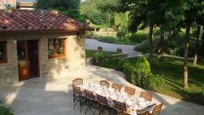 Ein Steinhaus mit Ziegeldach, eine Terrasse mit Esstisch und ein Garten mit Bäumen und Sträuchern.