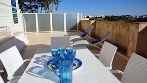 Une terrasse extérieure avec une table blanche, de la verrerie bleue et des chaises en osier blanc.