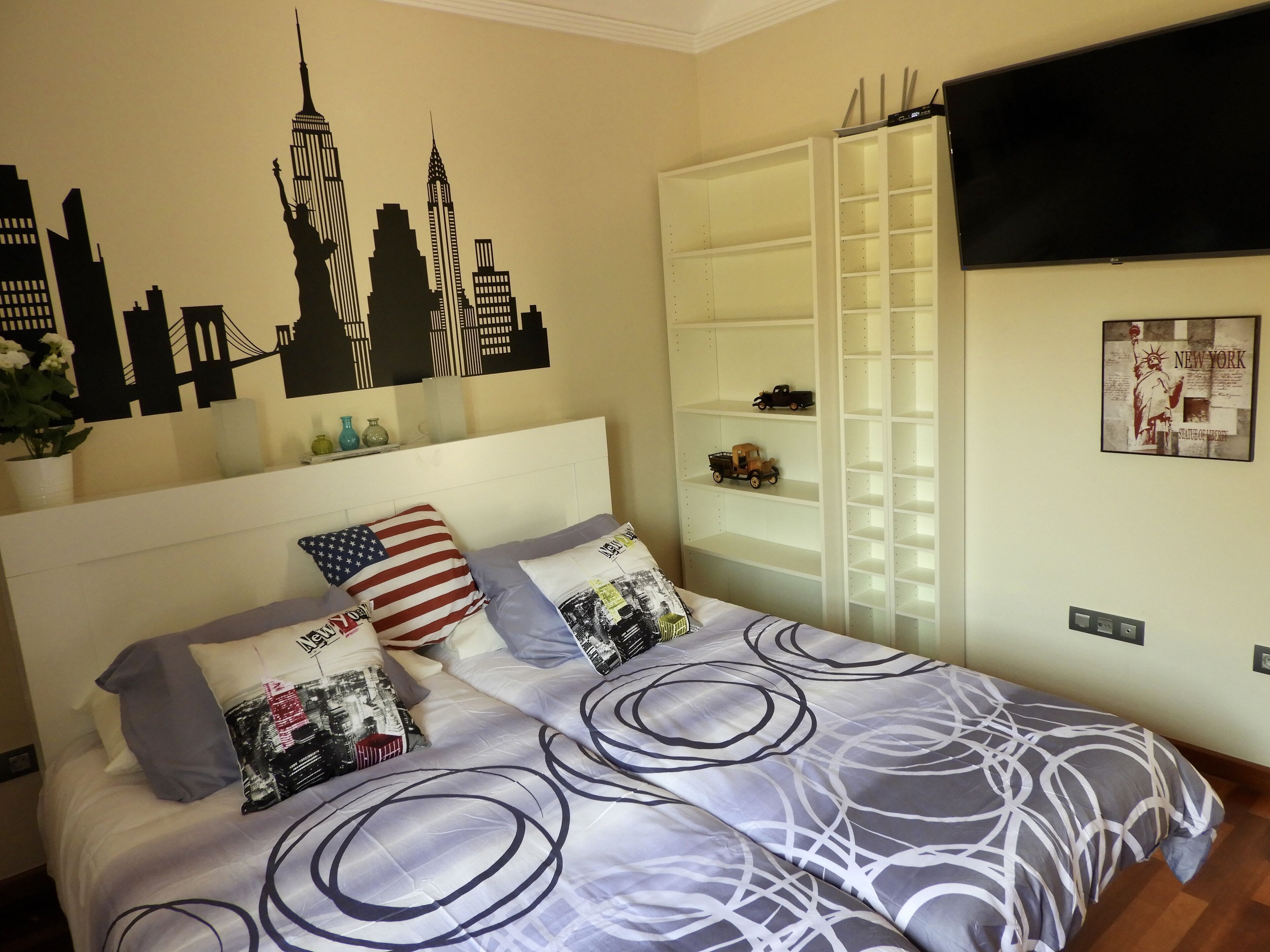 A bedroom with a bed featuring a city skyline mural, a white headboard, and a television mounted on the wall.