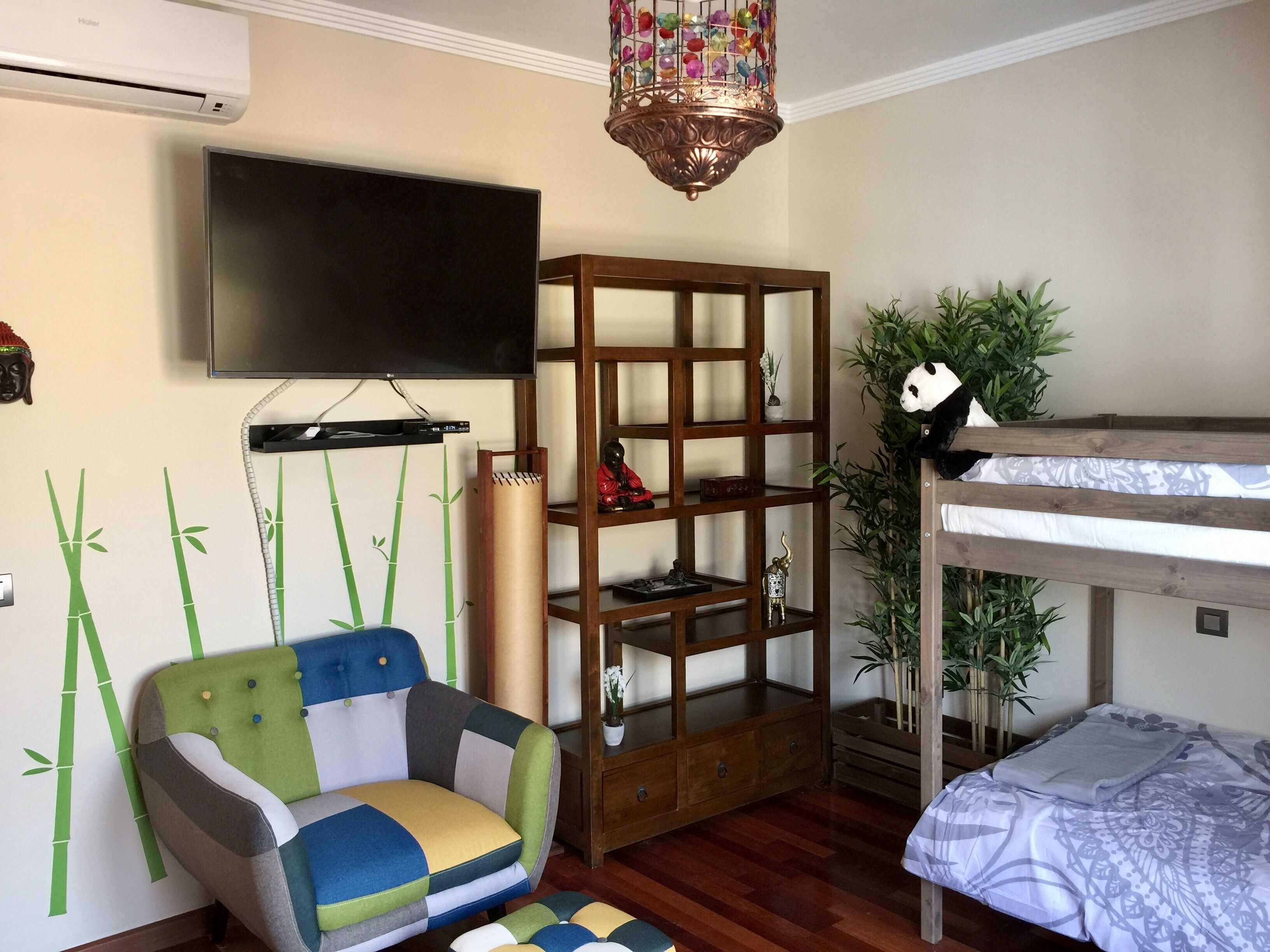 Una habitación con una cama litera, un televisor en la pared, una estantería de madera, un sillón colorido y adornos de bambú en las paredes.