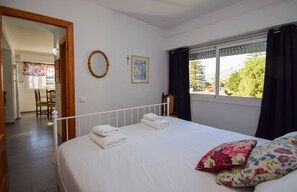 A bedroom with a bed, two pillows, a window with curtains, a wall-mounted picture, and a door leading to another room.