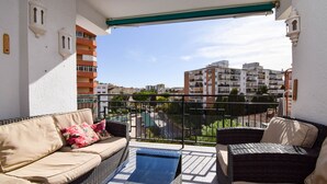 A balcony with a sofa and wicker chairs, a blue mat, and a view of buildings in the distance.