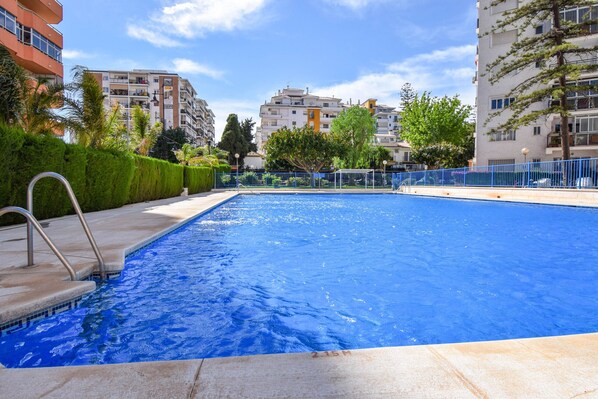 Una piscina con una escalera metálica, rodeada de una valla azul, y edificios al fondo.