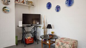 A living room with a flat-screen TV, a small round table with a lamp, a wicker basket, and a floral ottoman.