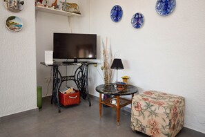 Una sala de estar con un televisor de pantalla plana, una mesita redonda con una lámpara, una cesta de mimbre y un reposapiés estampado con flores.