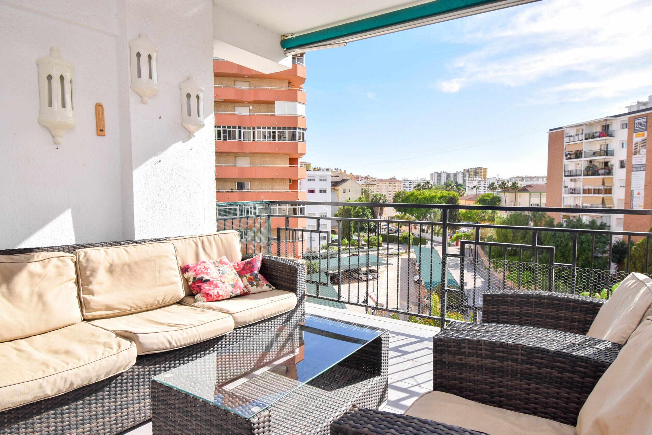 A balcony with wicker furniture, a glass table, and a view of a swimming pool and buildings.