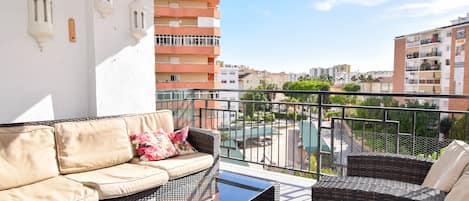 A balcony with wicker furniture, a glass table, and a view of a swimming pool and buildings.