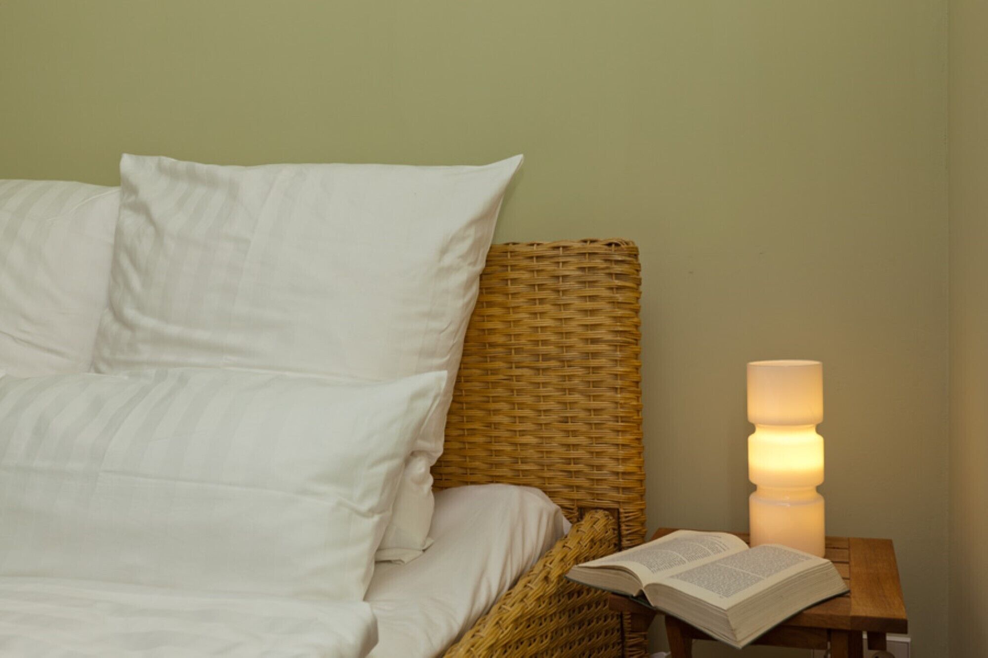 Un lit bien fait, avec du linge de lit blanc, une tête de lit en osier et une table de chevet sur laquelle se trouve une lampe allumée et un livre ouvert.