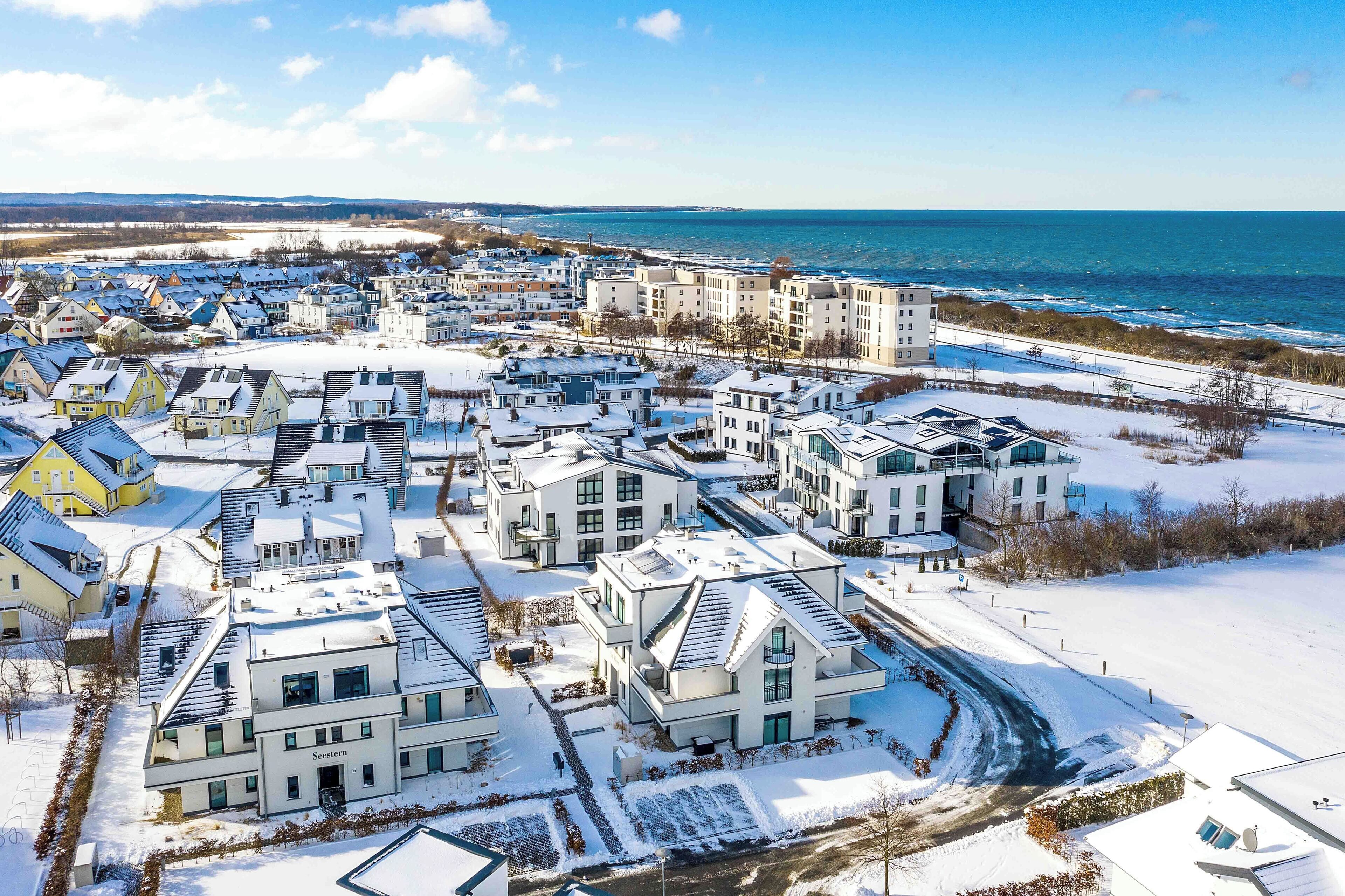 A snow-covered residential area with modern buildings, a clear view of the sea, and a road leading to the beach.