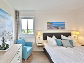 A hotel room with a bed, a chair, a desk, a nightstand, a lamp, a painting, and a window with curtains.