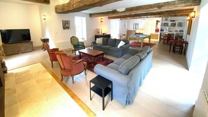 A spacious living area with a large sofa, a coffee table, and a television. There is a dining area with a pool table and chairs. The room has exposed wooden beams on the ceiling and a large window providing natural light.