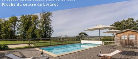 A swimming pool with wooden deck chairs and umbrellas, a wooden cabin, and a view of the countryside.