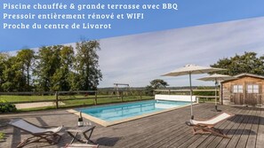 A swimming pool with wooden deck chairs and umbrellas, a wooden cabin, and a view of the countryside.