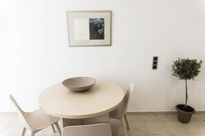 A minimalist dining area with a round table, two chairs, a bowl, a framed picture, a potted plant, and a wall-mounted switch. - Reflection of neutral warm `Urban Cocoa`` (Rethymno)