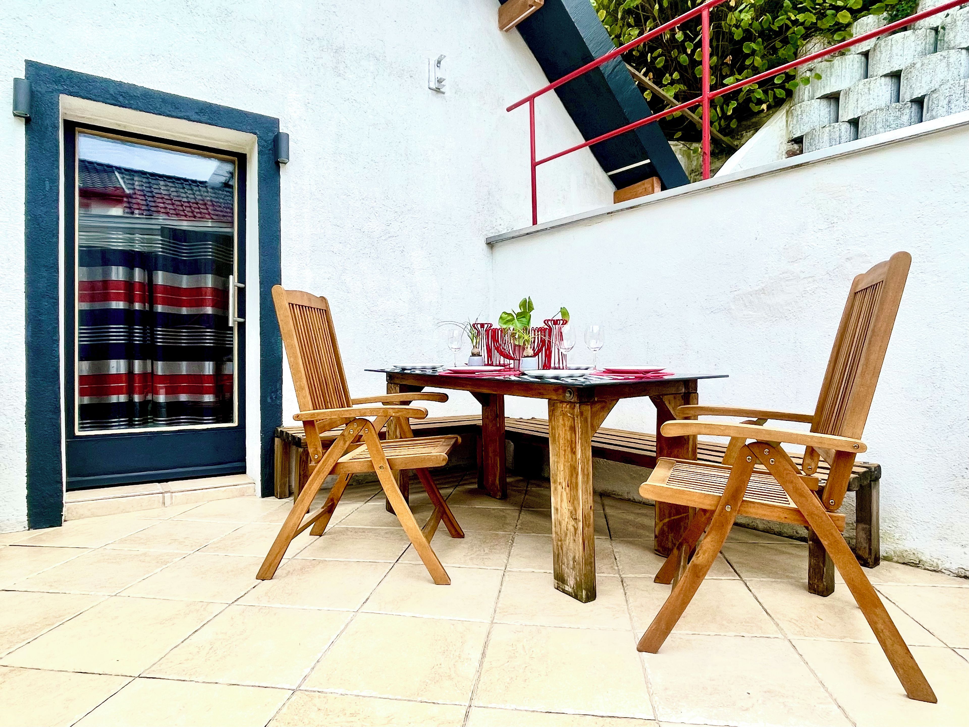 Un patio al aire libre con una mesa y cuatro sillas de madera, una barandilla roja y una ventana con cortina a rayas.