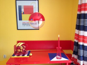 A red table with a fruit basket, a candle, and a framed picture on the wall.
