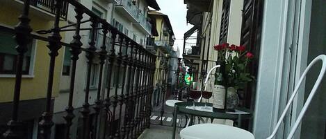 Un estrecho pasillo exterior con una cerca metálica, una pequeña mesa con sillas y un jarrón con flores.