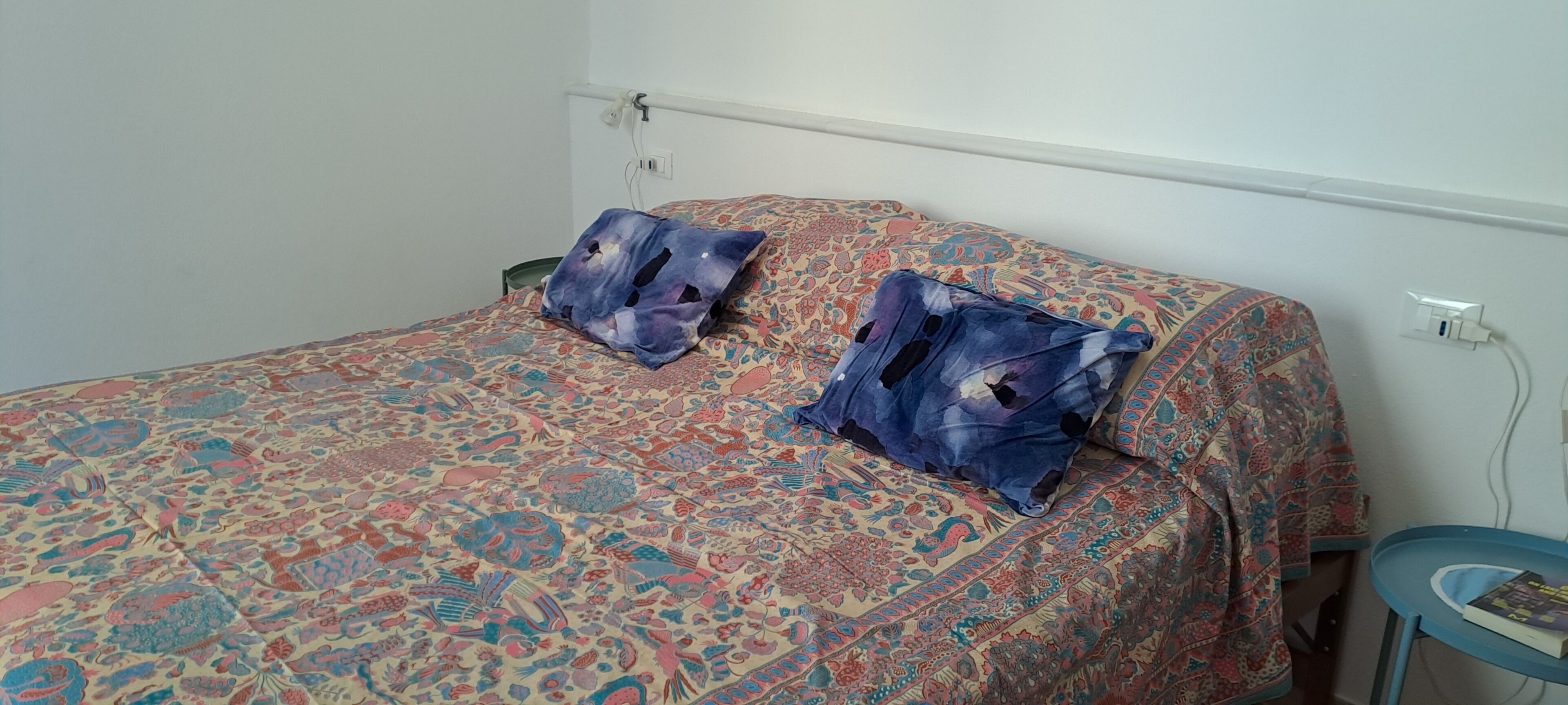 A bed with patterned bedding and two pillows, a bedside table with a book, and a power outlet on the wall.