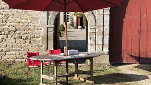Een rustieke buitenruimte met een houten tafel en rode stoelen, een parasol en een stenen muur op de achtergrond.