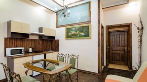 A compact kitchen with a microwave, kettle, and wooden cabinets. There is a dining table with chairs and a sofa in the living area. A painting hangs on the wall, and a wooden door is visible. - Apart-Hotel Kraft (Lviv)