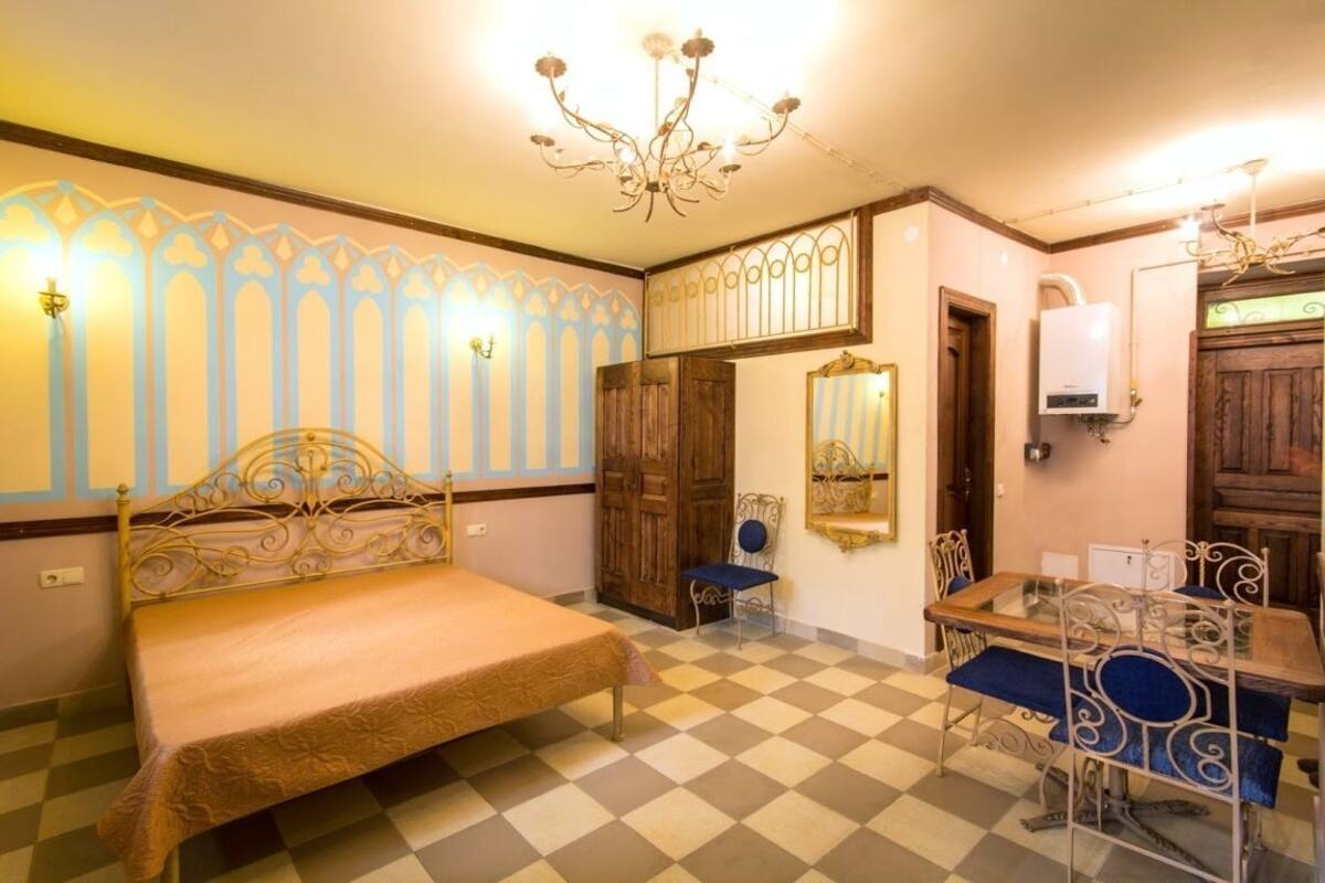 A room with a bed, a table with chairs, a chandelier, and a mirror.