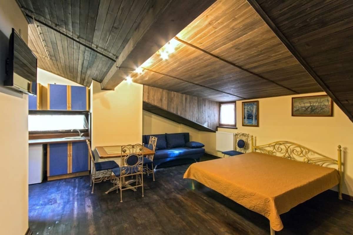 A compact room with a bed, a small table, and a sofa. There is a kitchenette with cabinets and a sink. The room has a sloped ceiling and wooden beams.
