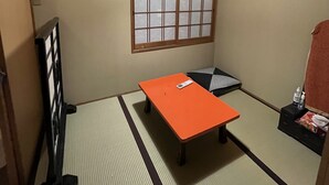 Una habitación con suelo de tatami, una mesa baja, un futón, una ventana con diseño de celosía y un ventilador de techo.