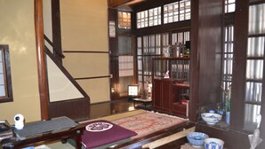 Una habitación tradicional japonesa con suelo de tatami, un escritorio de madera bajo, un futón de tatami con un diseño circular y una estantería de madera con varios objetos.