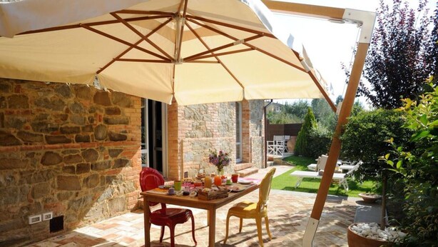 A stone patio with a table and chairs under a large umbrella, surrounded by greenery and a stone wall. - Villa Ventotto by Paola Orazioli (Arezzo)
