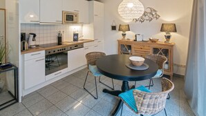 Een moderne keuken met witte kasten, een centraal eiland met een ronde tafel, en een eethoek met rieten stoelen.