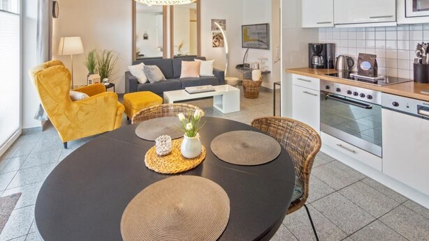 Een moderne woonkamer met een ronde eettafel, gele fauteuils, een bank en een keuken met een fornuis en een magnetron.