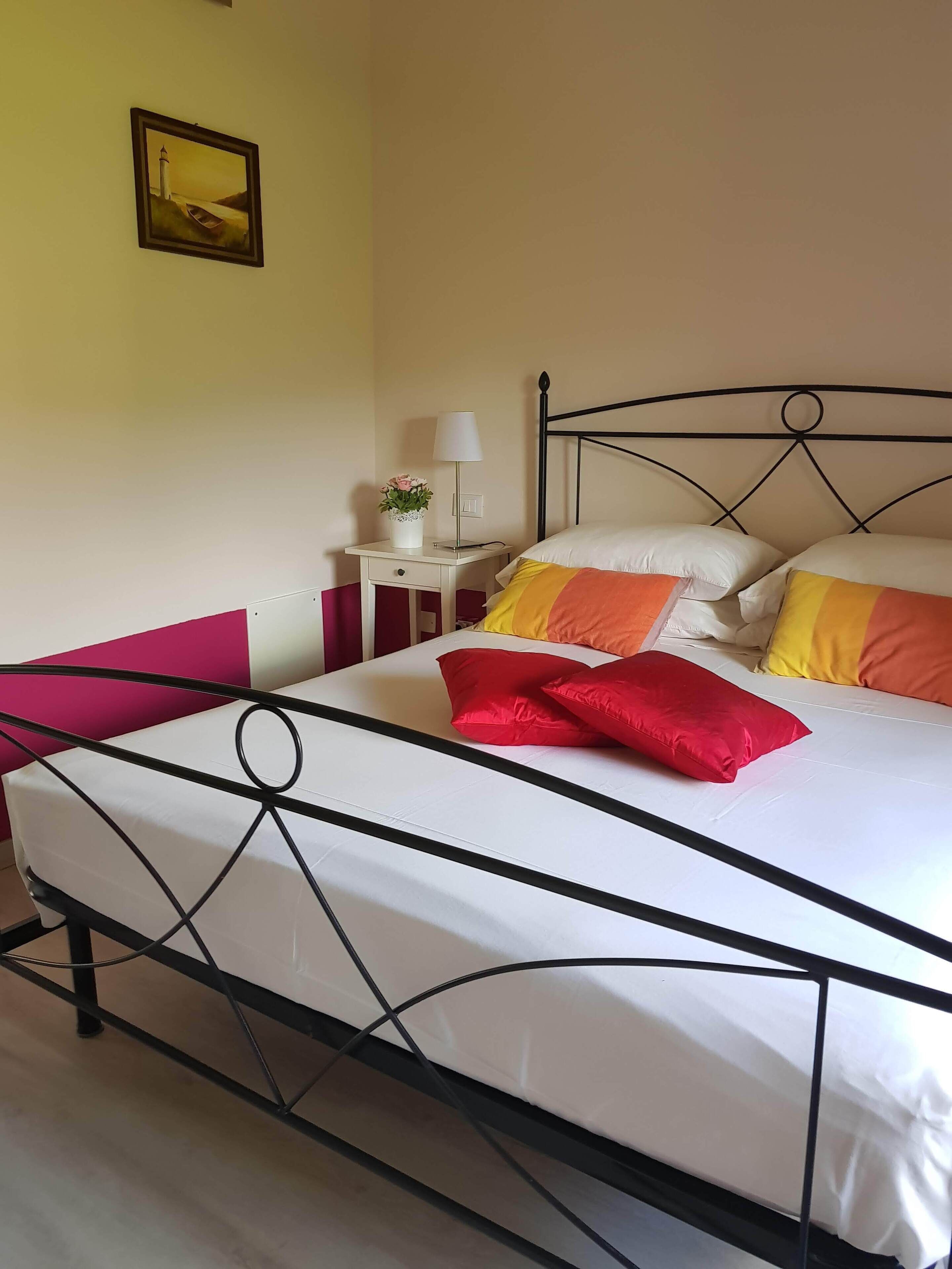 A bed with a metal frame, a bedside table with a lamp and a vase of flowers, and a painting on the wall.