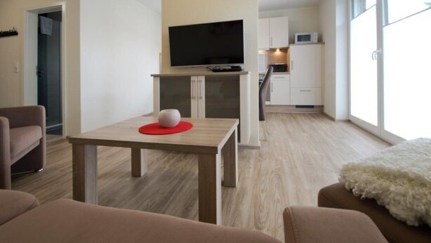 Een moderne woonkamer met een flatscreen televisie, een klein tafeltje met een rode onderzetter en een zacht wit tapijt.