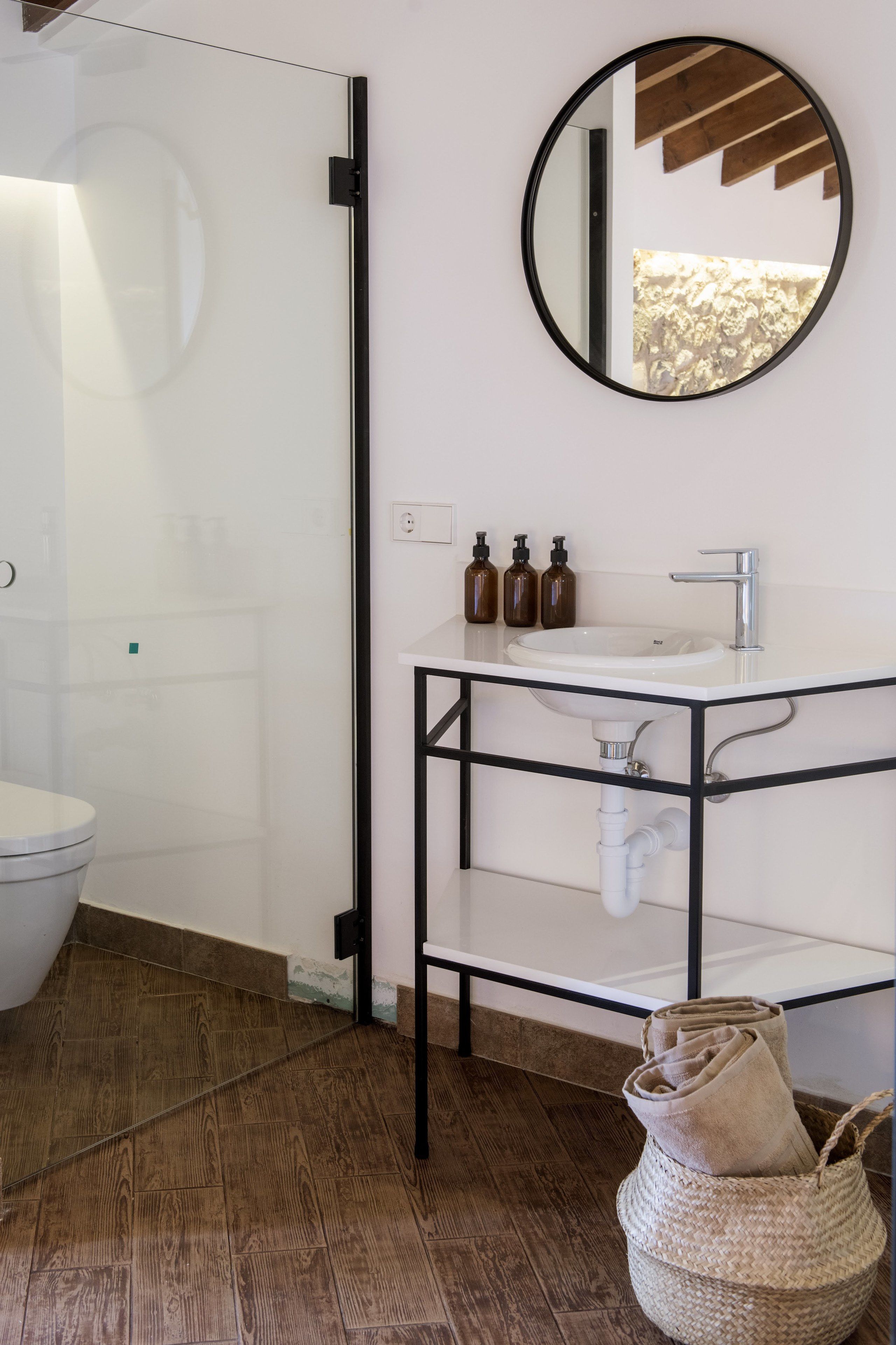 Un baño con mampara de vidrio, un lavabo blanco con estructura metálica negra y un espejo redondo en la pared.