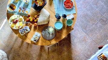 A round wooden table with various food items and utensils, surrounded by a tiled floor and a potted plant.