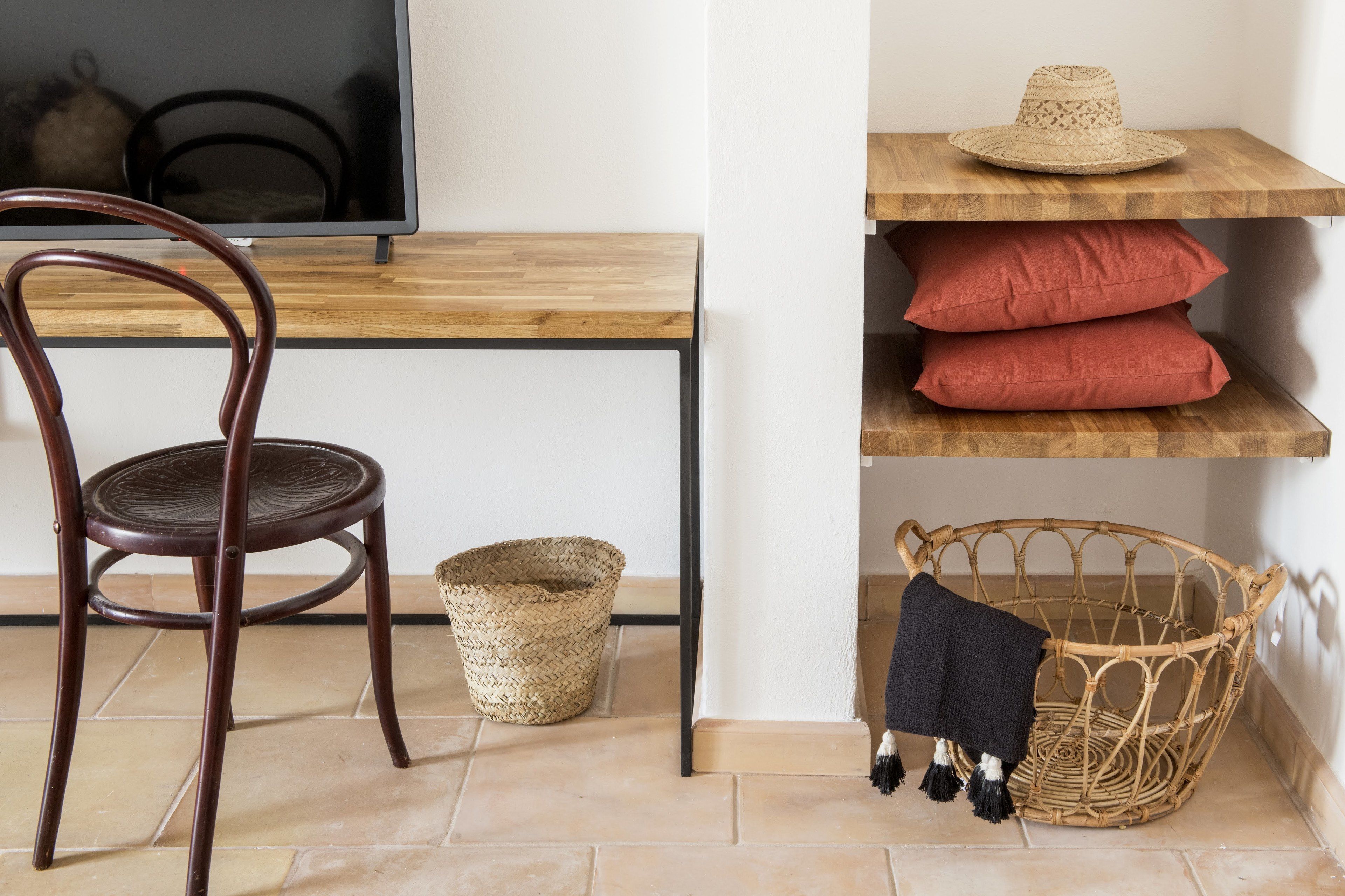 Un escritorio de madera con una cesta tejida y una silla, un televisor y estantes con sombreros y almohadas.
