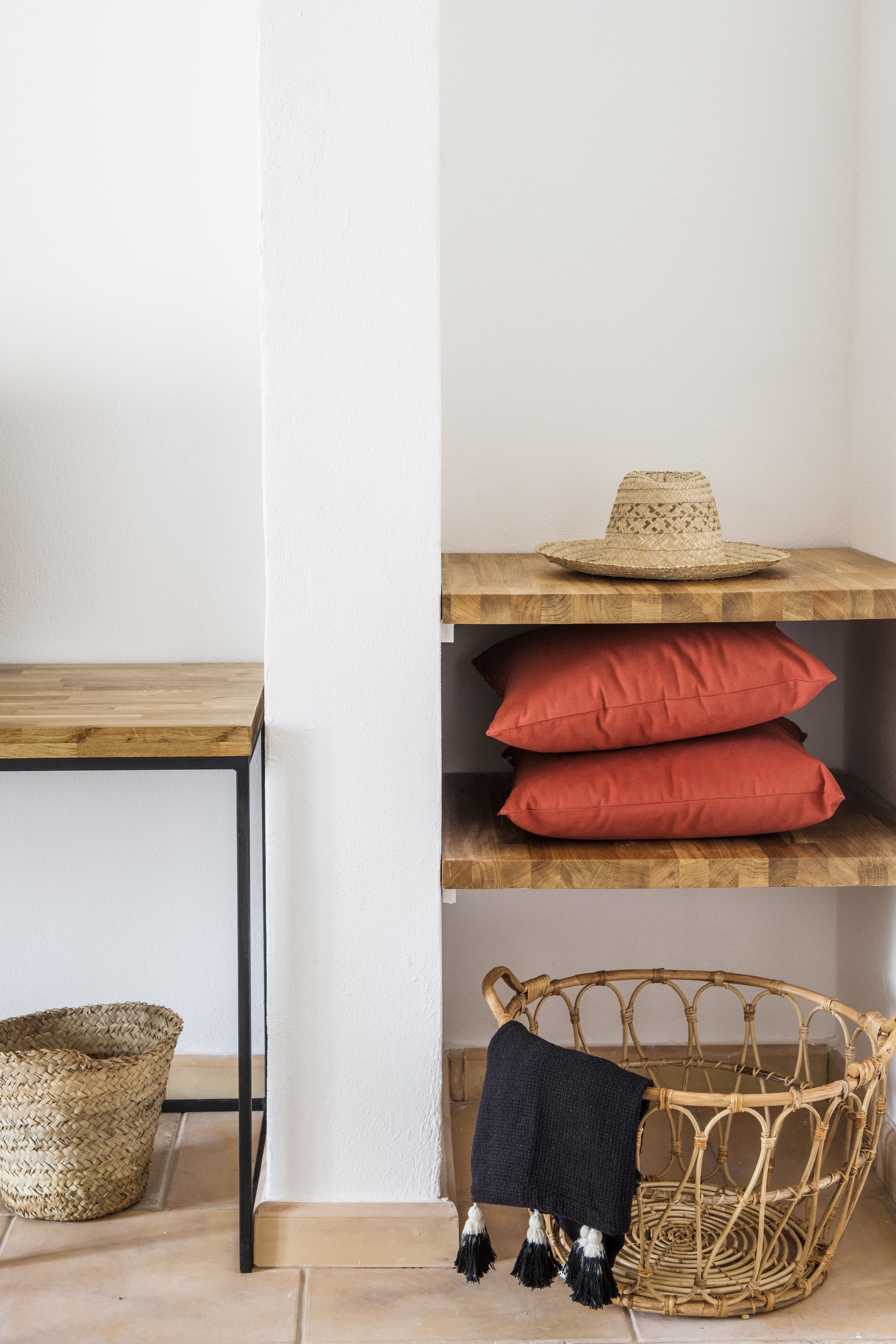 A corner of a room with a wooden table, a wicker chair, and a shelf with red cushions and a straw hat.