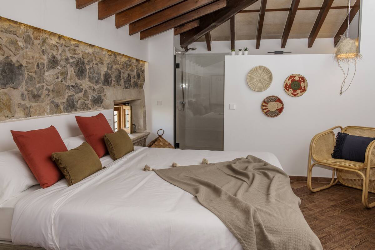 Un dormitorio con pared de piedra, cama con ropa de cama blanca y marrón, una silla de mimbre y una ducha de vidrio.